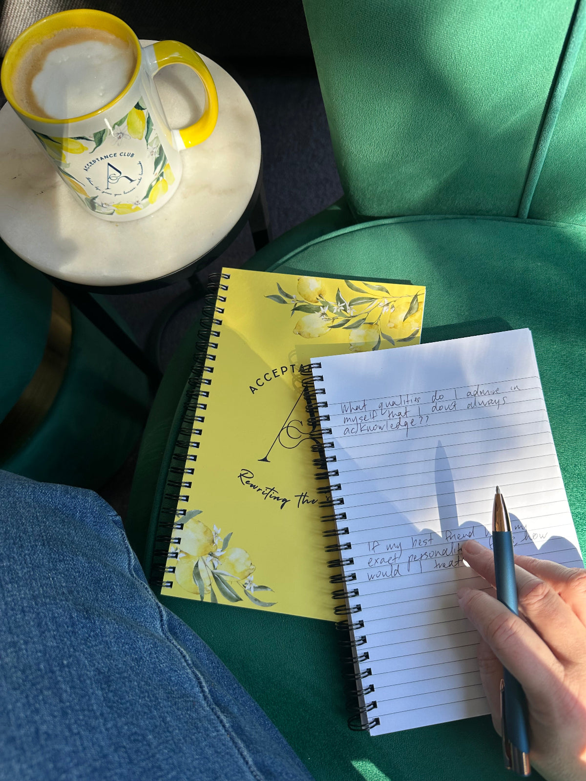 Person writing in a notebook with a yellow notepad and a cup of coffee on a green surface